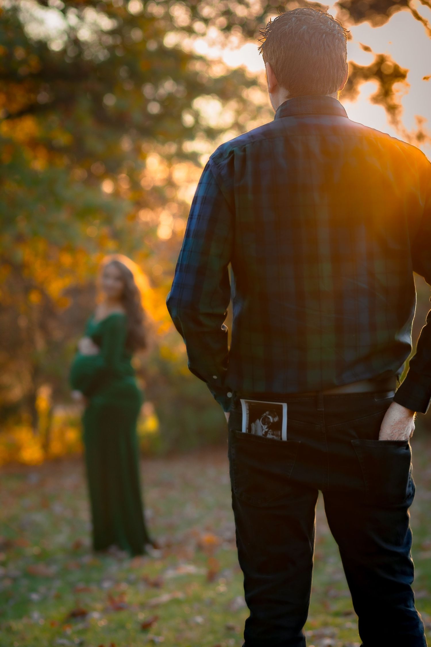 a man poses with the sonogram in his pocket while his pregnant wife's silhouette is in the background