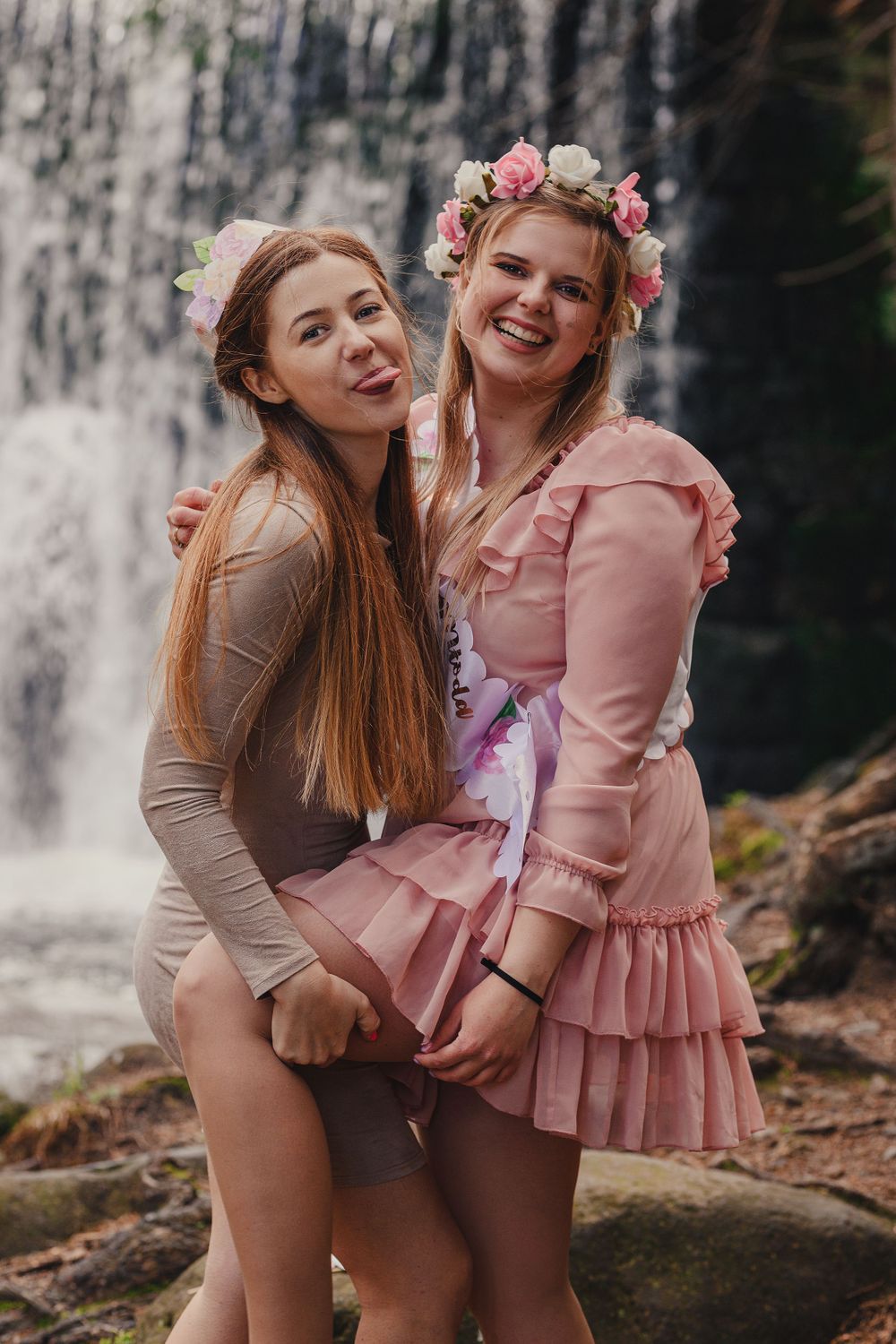 Friends posing together by a waterfall wearing pink and beige outfits in nature setting.