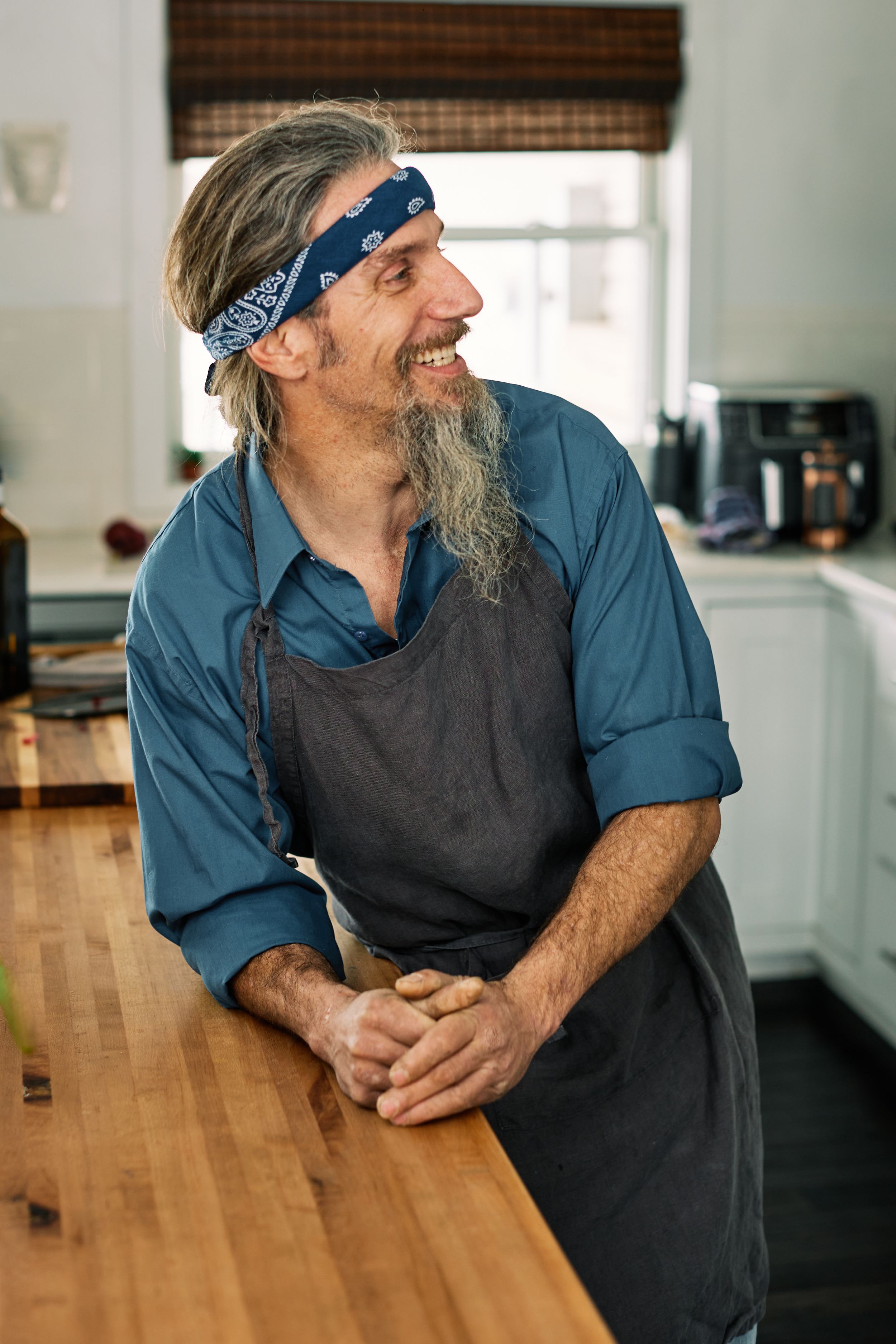 Chef in blue shirt and dark apron smiles while leaning on wooden counter in bright kitchen.