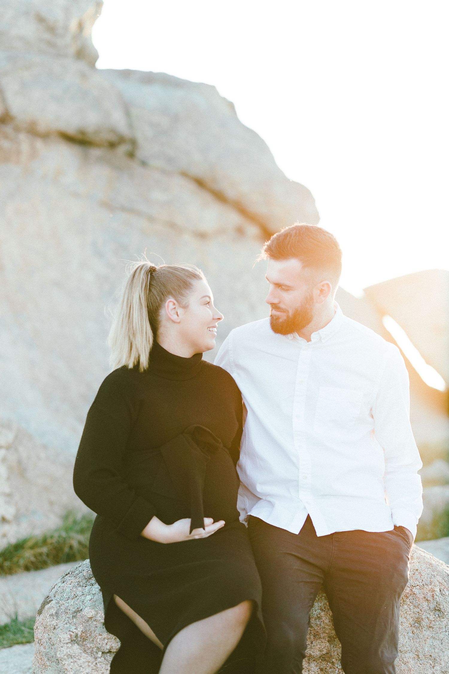 A couple shares a tender moment while sitting on rocky terrain at sunset.