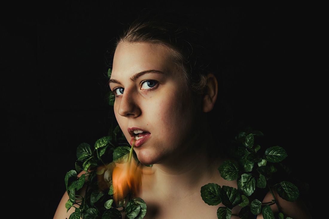 Dramatic close-up portrait surrounded by green leaves and orange lily against black background.