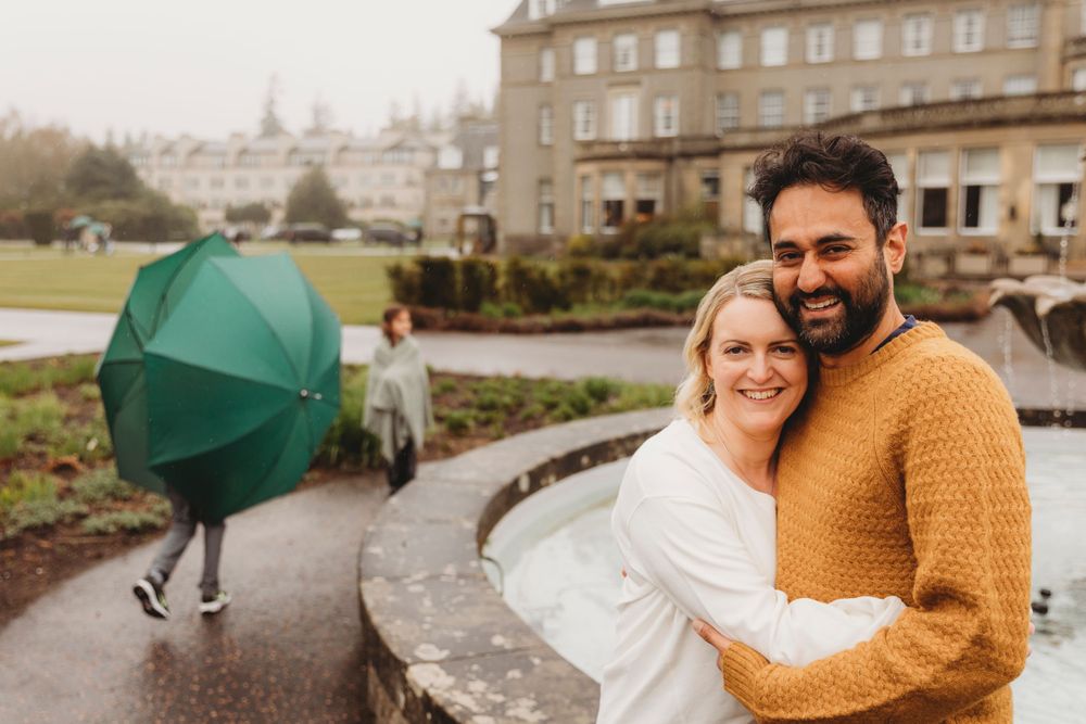A couple embraces and smiles on a rainy day with an emerald umbrella in the background.