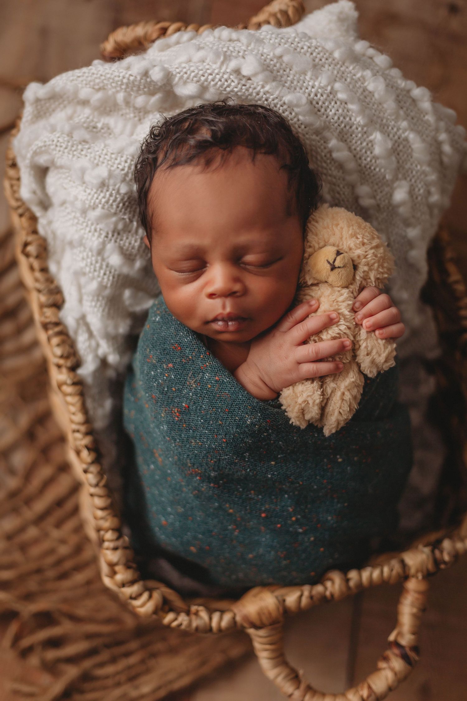A sleeping baby wrapped in teal swaddle holds teddy bear in basket with white blanket.