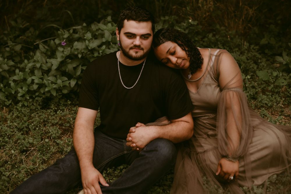 A couple in contrasting outfits sits close together against a backdrop of green leaves.