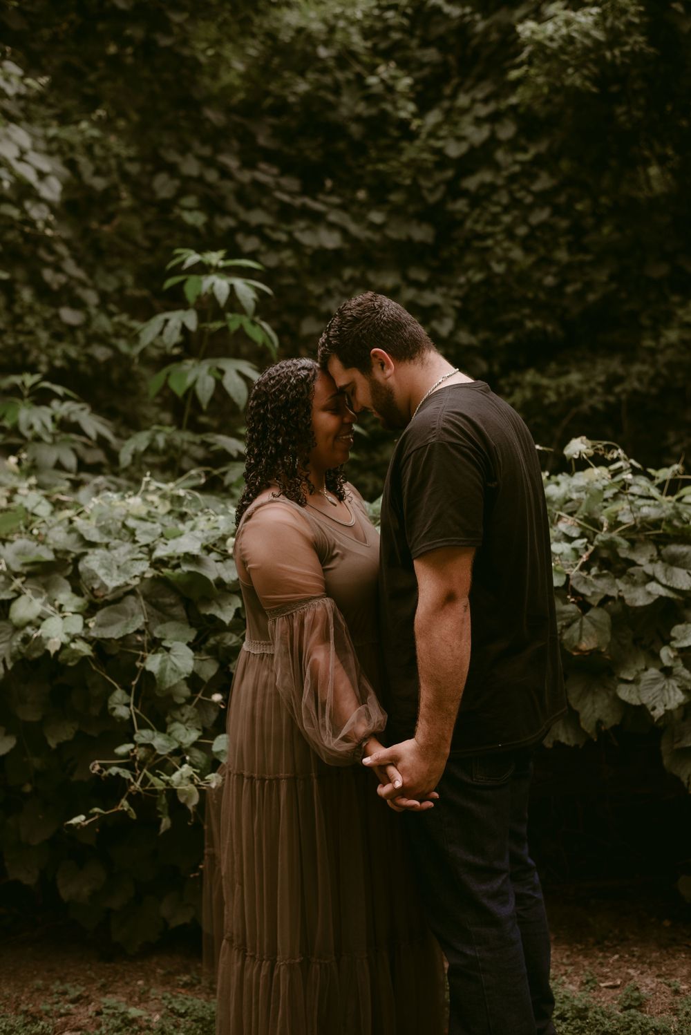 An intimate couple embraces while holding hands against a backdrop of lush green foliage.