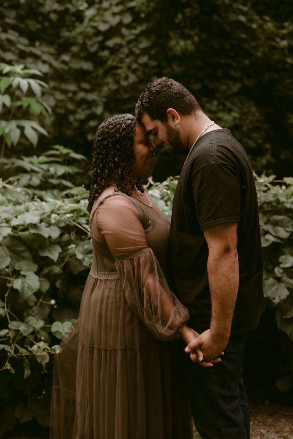 A romantic couple shares a tender forehead touch moment while holding hands in a forest setting.