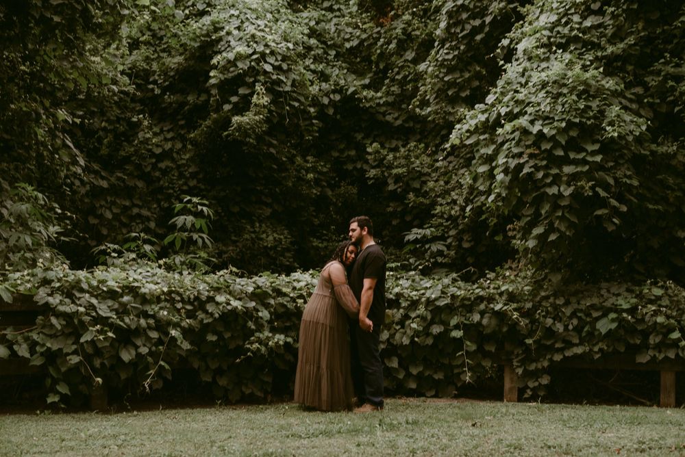 A couple embraces in a garden surrounded by green foliage during a moody outdoor photoshoot.