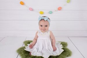 Baby in white dress sits on green furry rug under colorful hanging garland decorations.