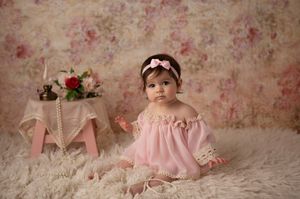 Baby in pink dress sitting on fur rug with floral vintage backdrop.