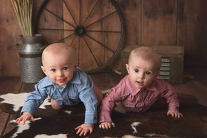 Two babies in matching outfits crawl on cowhide rug with rustic barn wood backdrop.