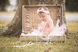 Baby in tulle dress and pearls poses in vintage suitcase outdoors near tree.
