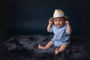 Baby in blue romper and straw hat sits on dark furry rug with black backdrop.