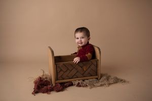 Baby in red sweater sits in wooden crate with textured fabric backdrop.