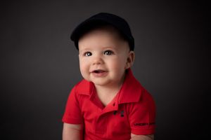 Baby wearing red polo shirt and navy cap against dark background.