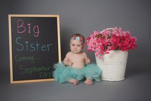 Baby in turquoise tutu next to chalkboard announcement and pink flowers.