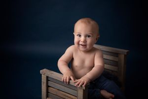 Baby sitting in wooden chair against dark blue studio backdrop.