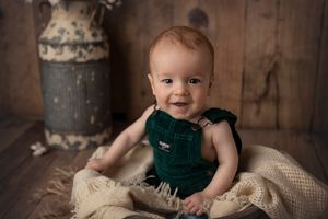 Baby in green overalls sitting among vintage props with rustic wooden background.