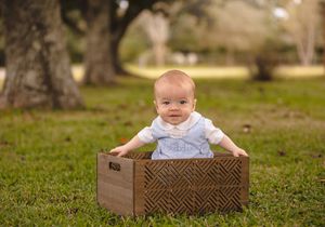 Baby sitting in wooden crate outdoors with trees in background.