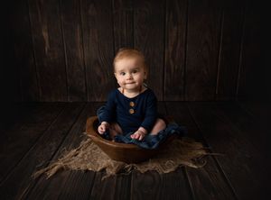 Baby in navy outfit sits in wooden bowl on rustic brown backdrop.