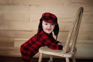 Baby wearing red plaid buffalo check outfit leaning on wooden chair.