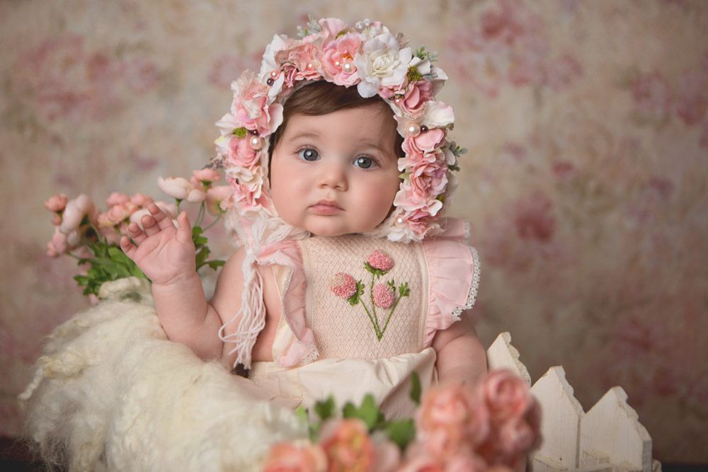 Baby in pink dress with floral bonnet against soft floral backdrop.