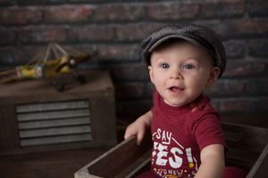 Baby in red shirt and newsboy cap against rustic brick background.