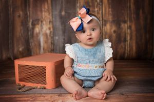 Baby in blue outfit with matching bow sitting by vintage orange radio.