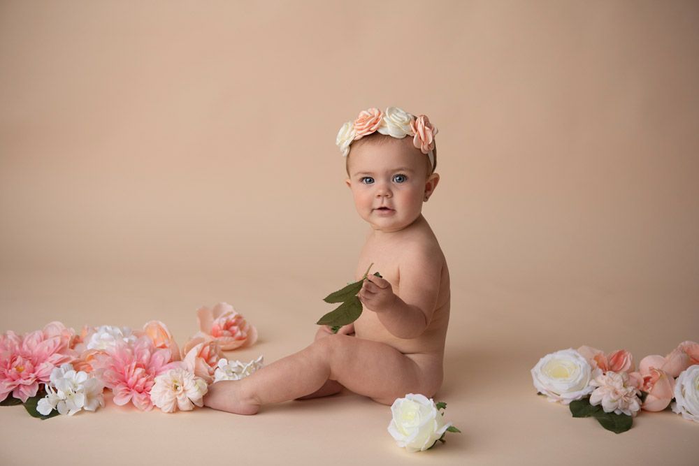 Baby wearing floral headband sitting on beige backdrop with pink flowers scattered around.