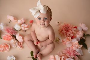 Baby with white bow headband surrounded by pink and peach flowers on beige background.
