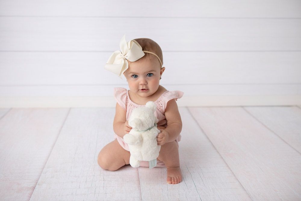 Baby wearing white dress and flower headband sitting on white wooden floor.