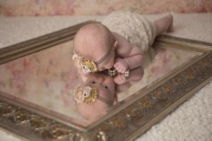 Baby in lace outfit lying on antique mirror with ornate gold frame.
