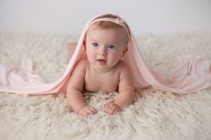Baby lying on white fluffy rug with pink blanket draped overhead.