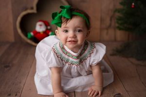 Baby in white smocked dress with green bow on wooden floor with Christmas decorations.