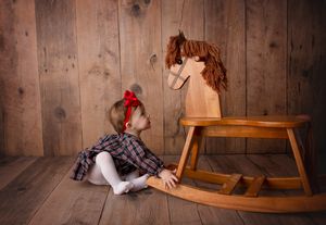 Small child in plaid dress reaching for wooden rocking horse against rustic wall.