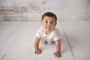 Baby in white onesie crawling on white wooden floor during photo session.