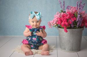 Baby in floral outfit sits beside galvanized bucket with pink flowers.