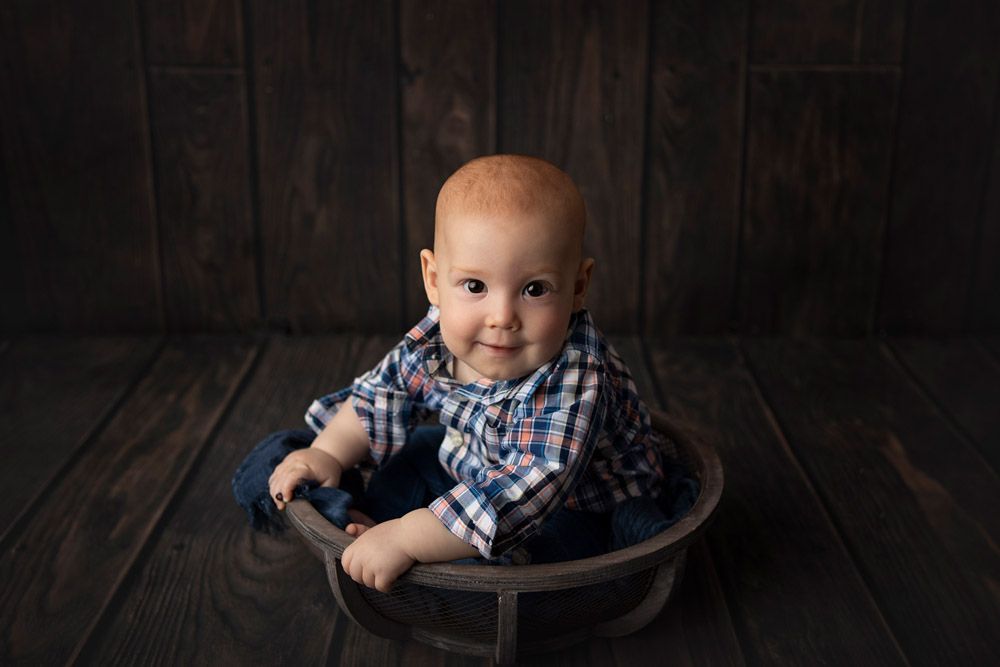 Baby in plaid shirt sitting in wooden bowl against dark backdrop.