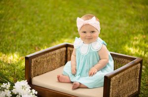 Baby in mint green dress sitting in wicker chair on grass with white flowers.