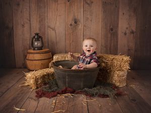 Baby sitting in rustic metal tub with hay bales and lantern in barn setting.