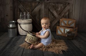 Baby in overalls sitting with vintage milk jug in rustic barn setting.
