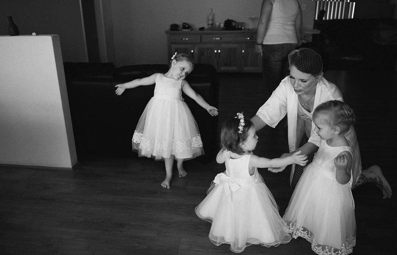 Young children in white tutus dance while an adult helps them in a casual indoor setting.