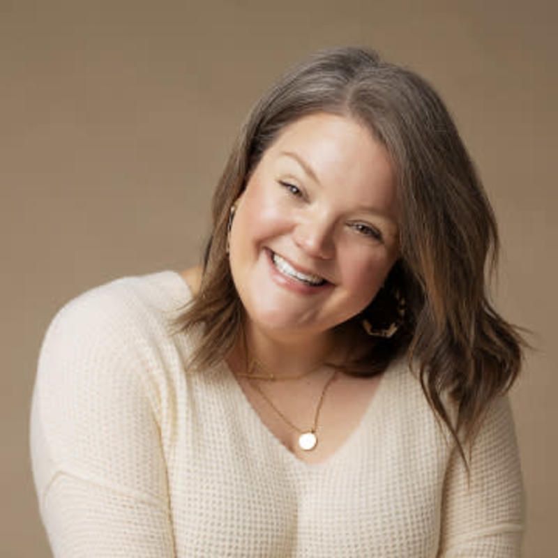 Smiling woman with shoulder-length hair wearing a cream sweater and delicate necklace against a warm beige background.
