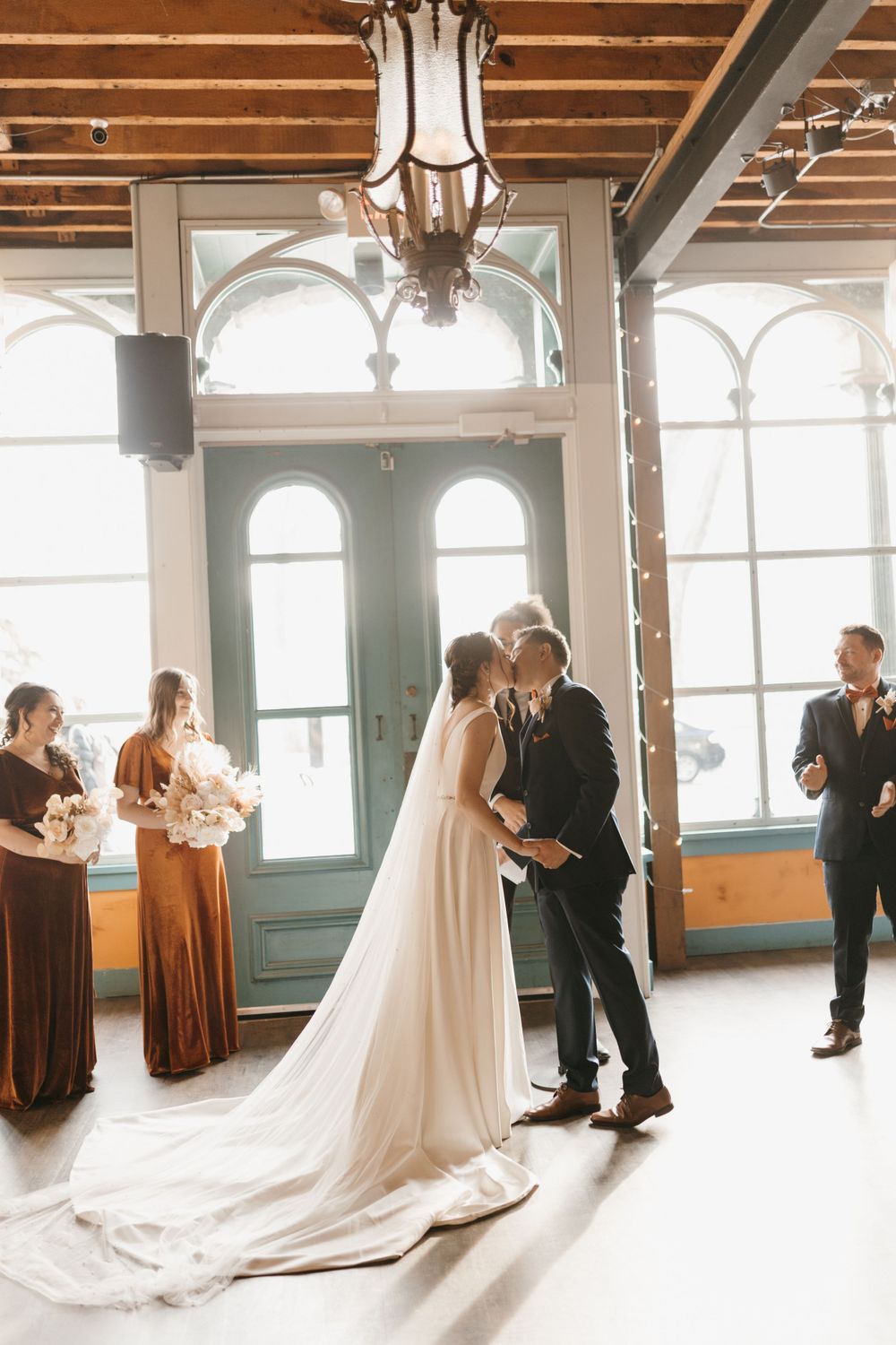 Newlyweds share first kiss at indoor wedding venue with bridal party in burnt orange.