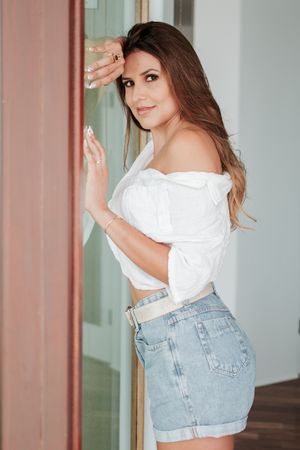 Woman in white crop top and denim shorts leans against wall in casual pose.