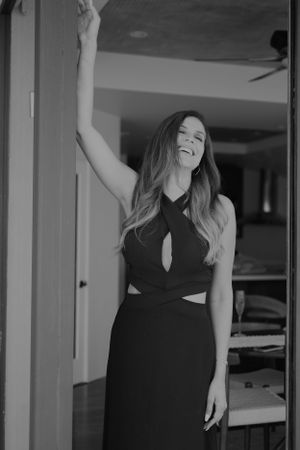 Woman in elegant black dress poses joyfully with raised arm in indoor setting.