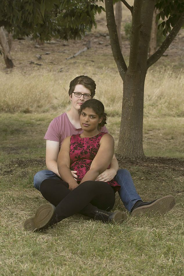 A couple sits intimately under a tree in a grassy field wearing pink and red floral outfits.