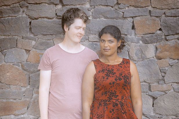 A couple poses against a rustic stone wall, wearing coordinating pink and red patterned outfits.