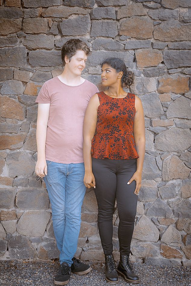 A couple stands against a stone wall background holding hands in casual pink and red attire.