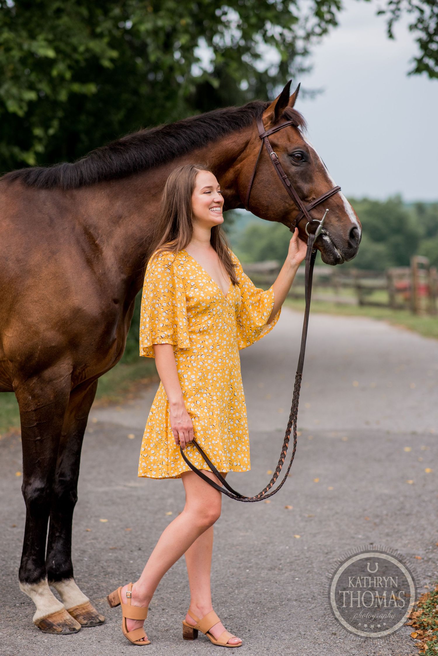 A bay horse and person in yellow floral dress pose together on a rural path.