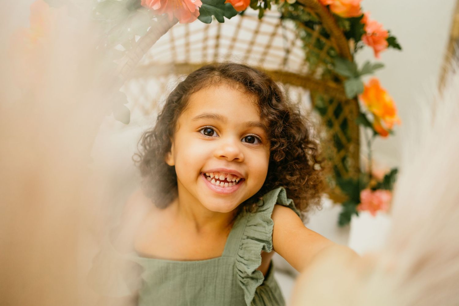 Adorable curly-haired toddler girl in green dress smiling brightly near a floral backdrop indoors.